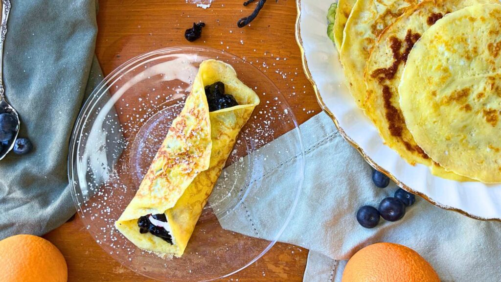 A crêpe with cream and blueberry filling on a glass plate. There are more crêpes off to the side.