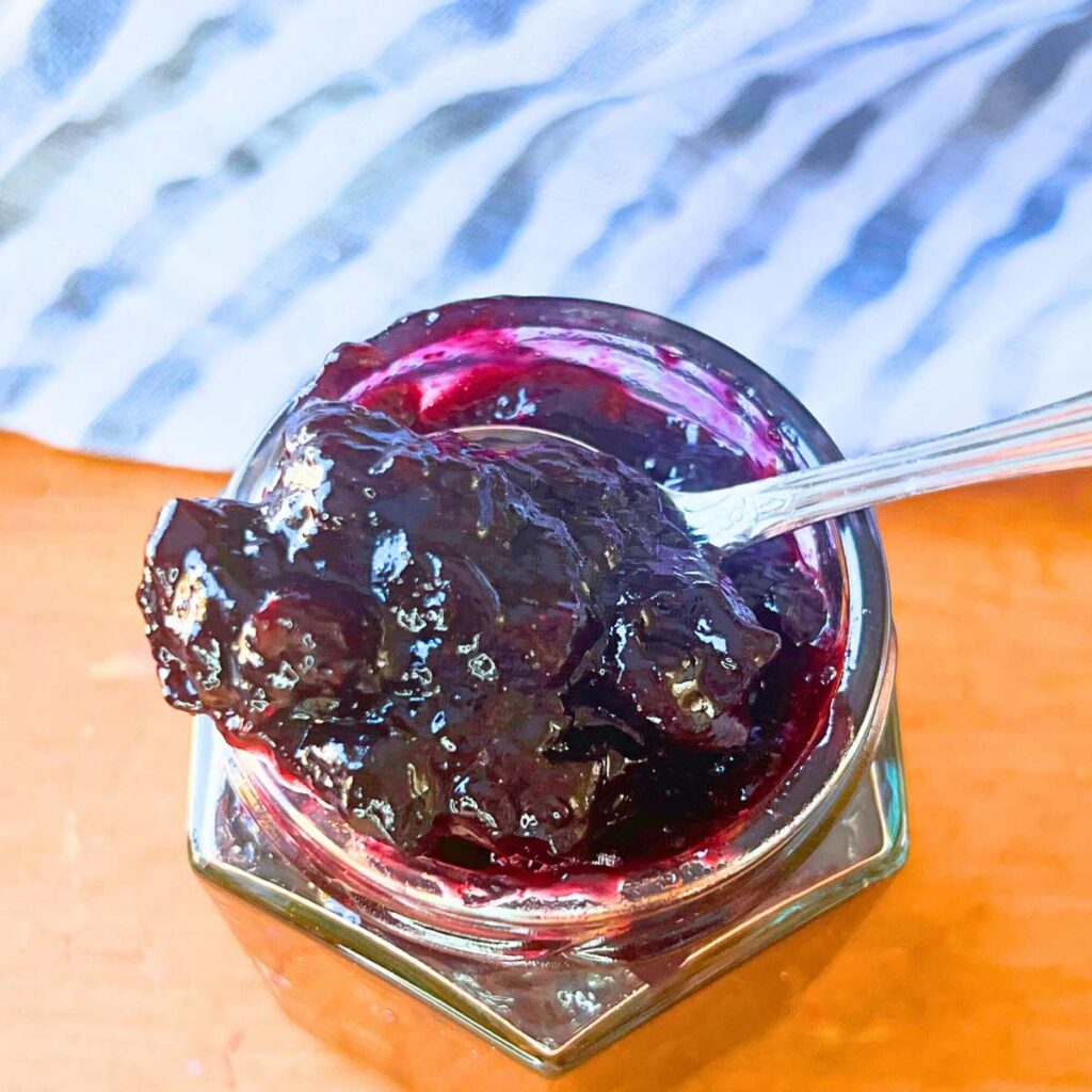 A glass jar full of blueberry sauce. There is a spoonful resting on top.