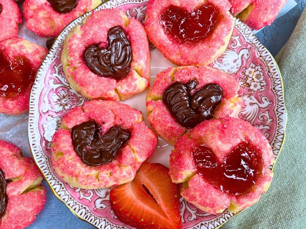 A pink plate full of thumbprint cookies coated in pink sugar. The heart shaped depression is filled with jam and chocolate.