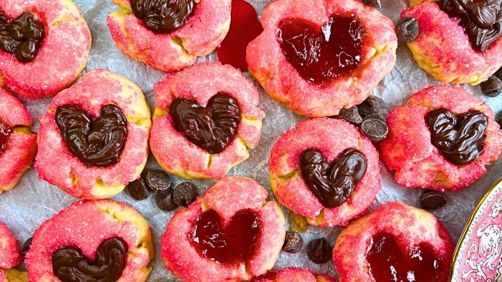 A table full of thumbprint cookies coated in pink sugar. The heart shaped depression is filled with jam and chocolate.