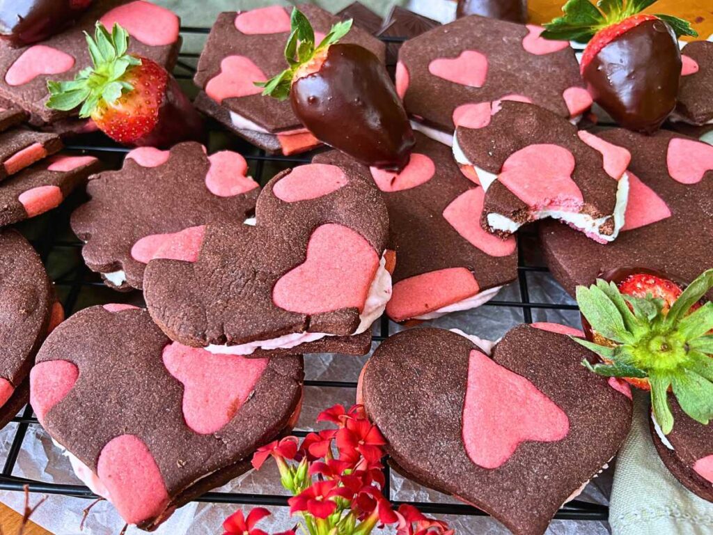 A collection of two-tone heart sandwich cookies surrounded by chocolate covered strawberries.