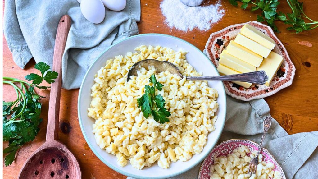 A large bowl with spaetzle noodles inside. There are butter, eggs, and flour in the background.