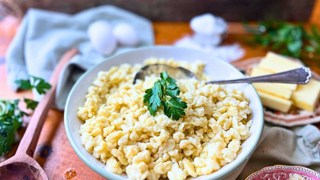 A large bowl of spaetzle noodles with parsley on top and a metal serving spoon.