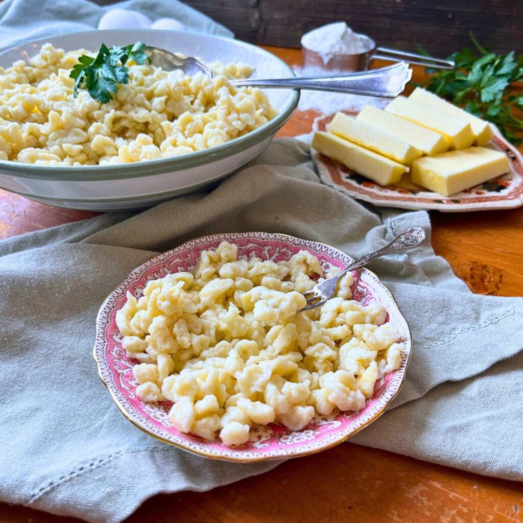 A small pink plate with homemade spaetzle noodles. There are more spaetzle in a large bowl in the background.