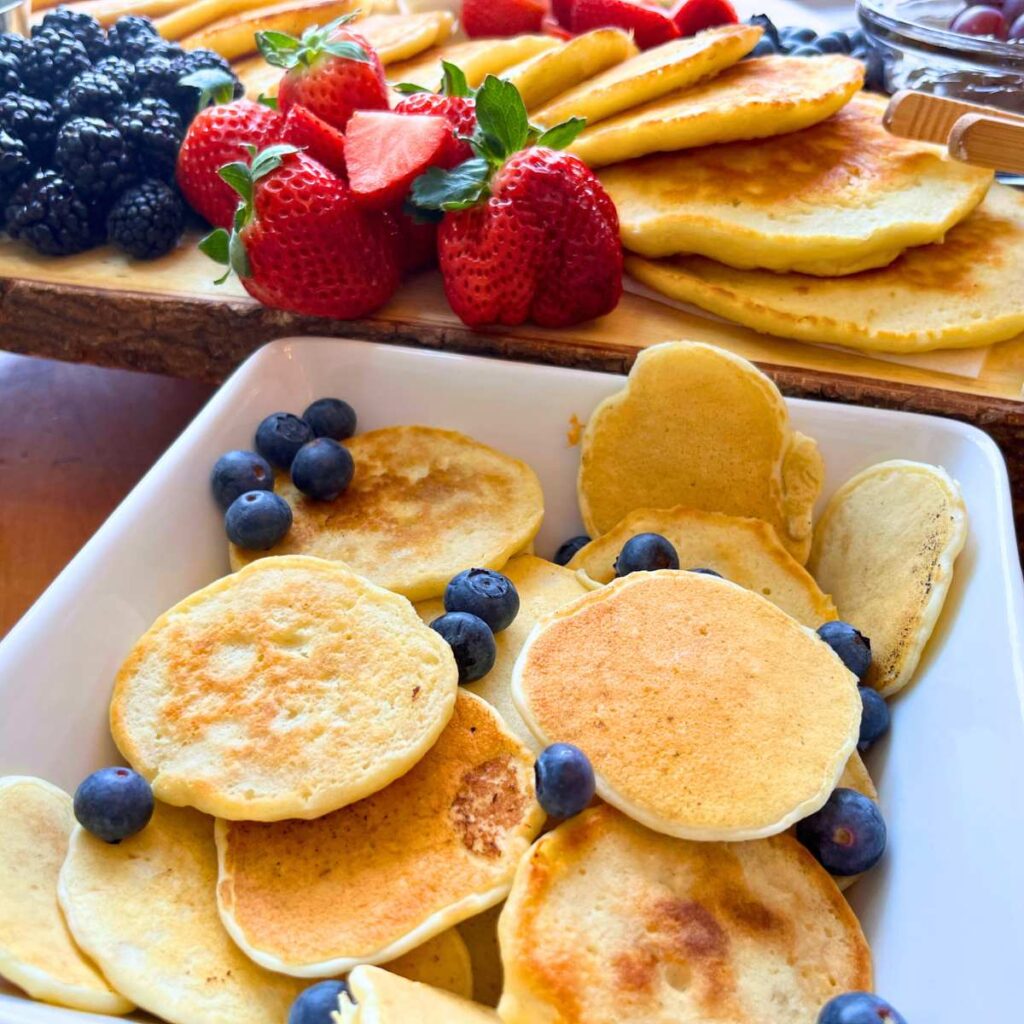 A white square bowl with mini pancakes and blueberries inside.