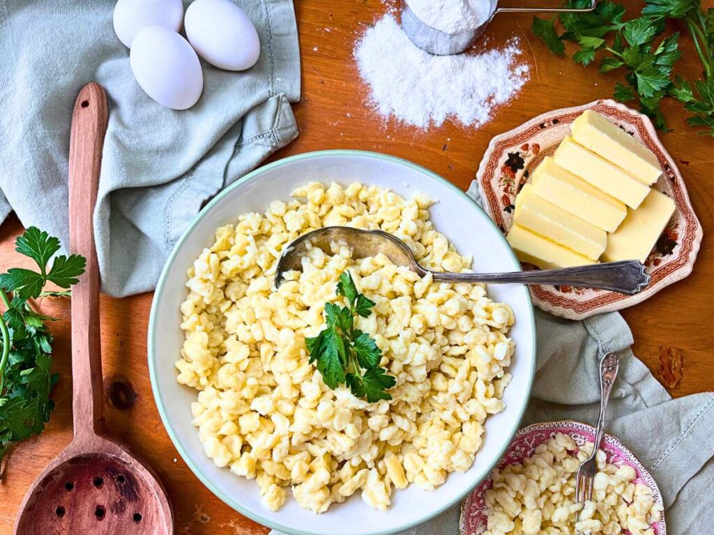 A bowl of spaetzle noodles in a large white bowl. There is a serving spoon in the pasta. There are eggs, flour, and butter in the background.