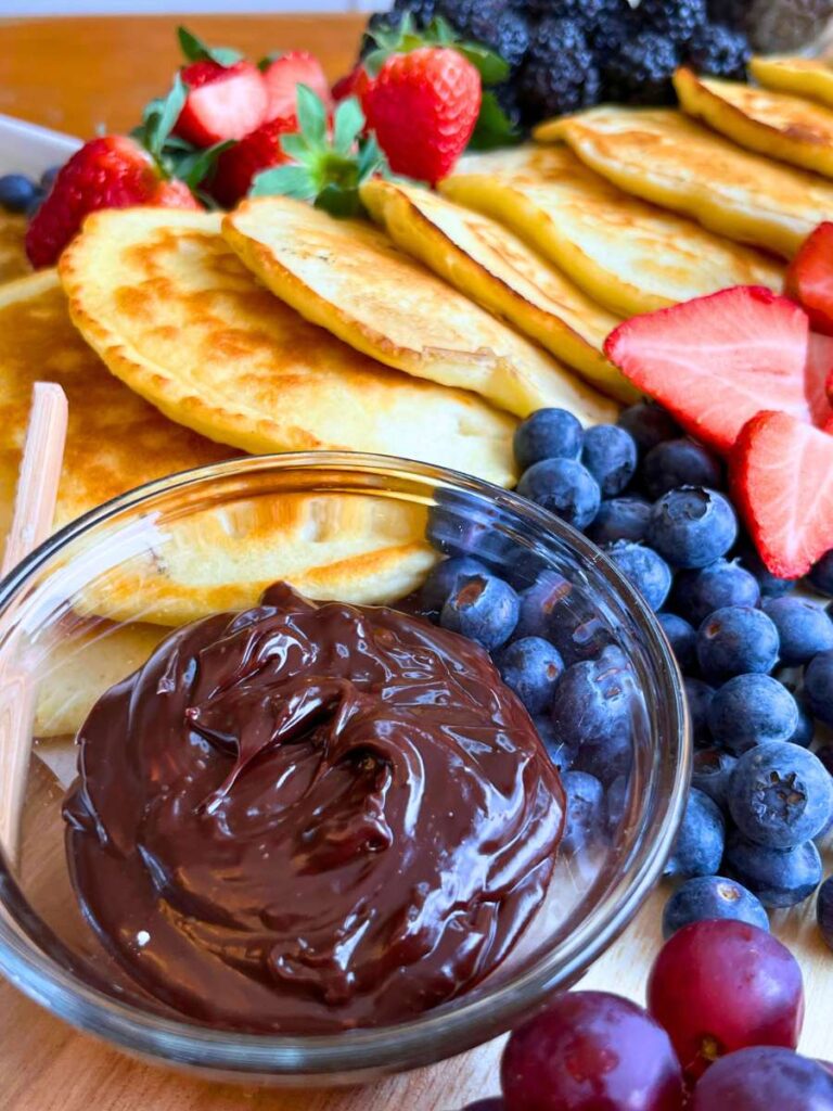 A glass bowl full of chocolate spread. There are pancakes and fresh fruit in the background.