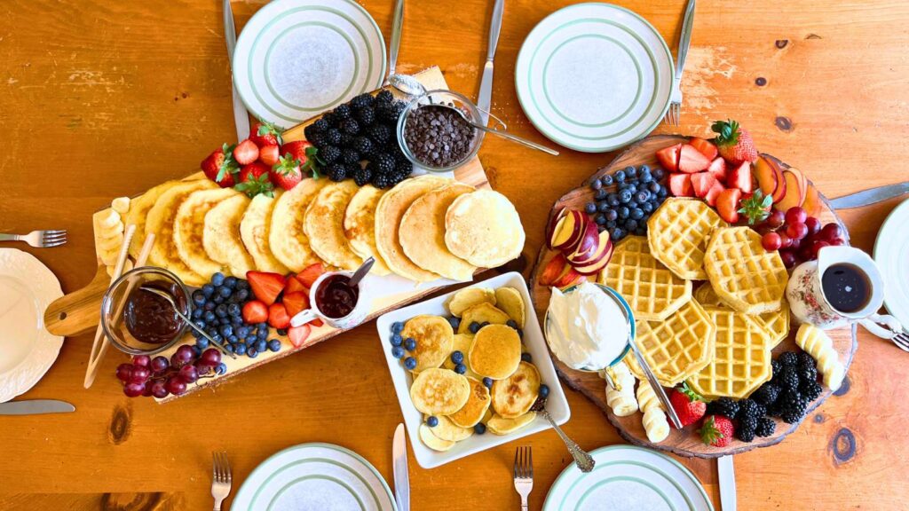 A wooden board with pancakes and fresh fruit. There is a board with waffles surrounded by fresh fruit and whipped cream. A large bowl with silver dollar pancakes with blueberries on top.