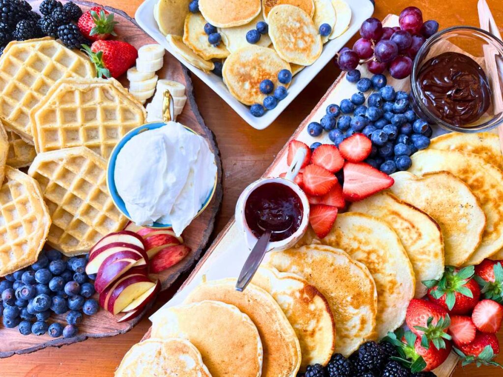 A board of pancakes and waffles surrounded by fresh fruit, whipped cream, and jam.