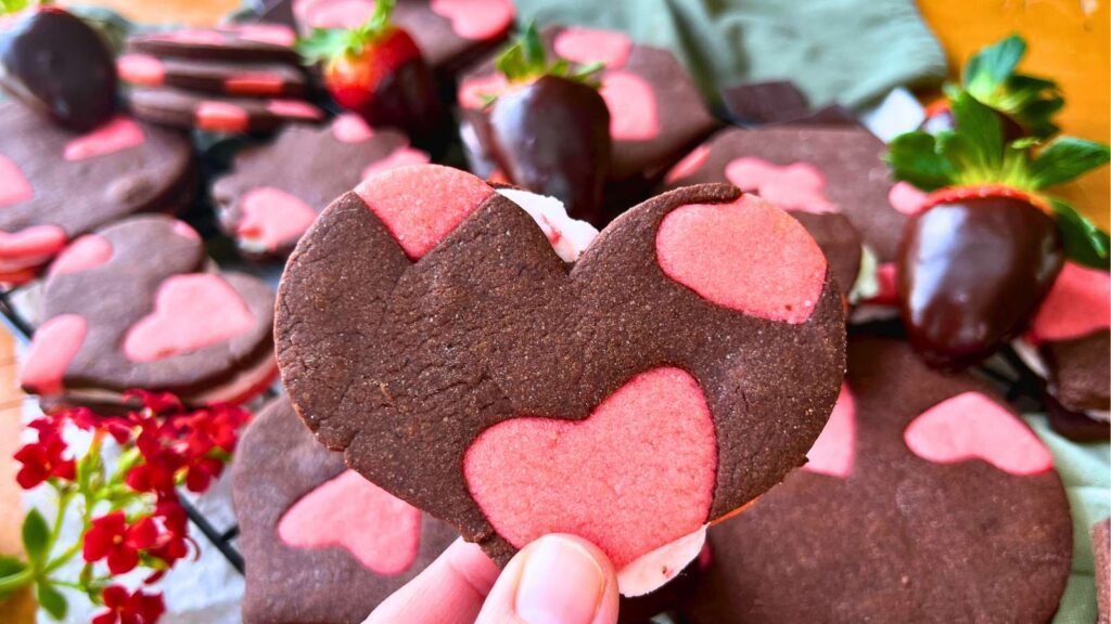 A woman holding two tone heart sandwich cookie. There is more in the background.