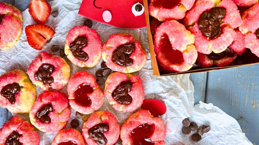 A table full of thumbprint cookies coated in pink sugar. The heart shaped depression is filled with jam and chocolate. Some are in a cardboard box.