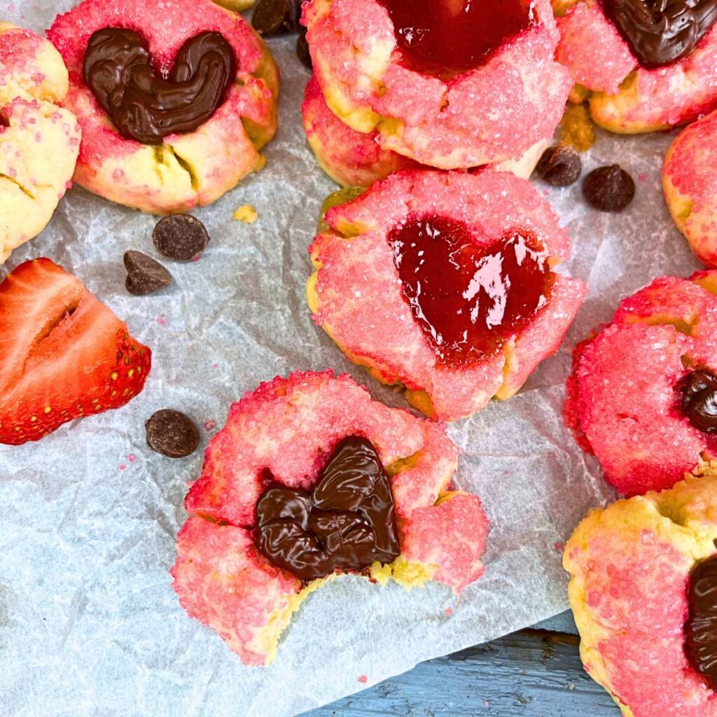 A table full of thumbprint cookies coated in pink sugar. The heart shaped depression is filled with jam and chocolate.