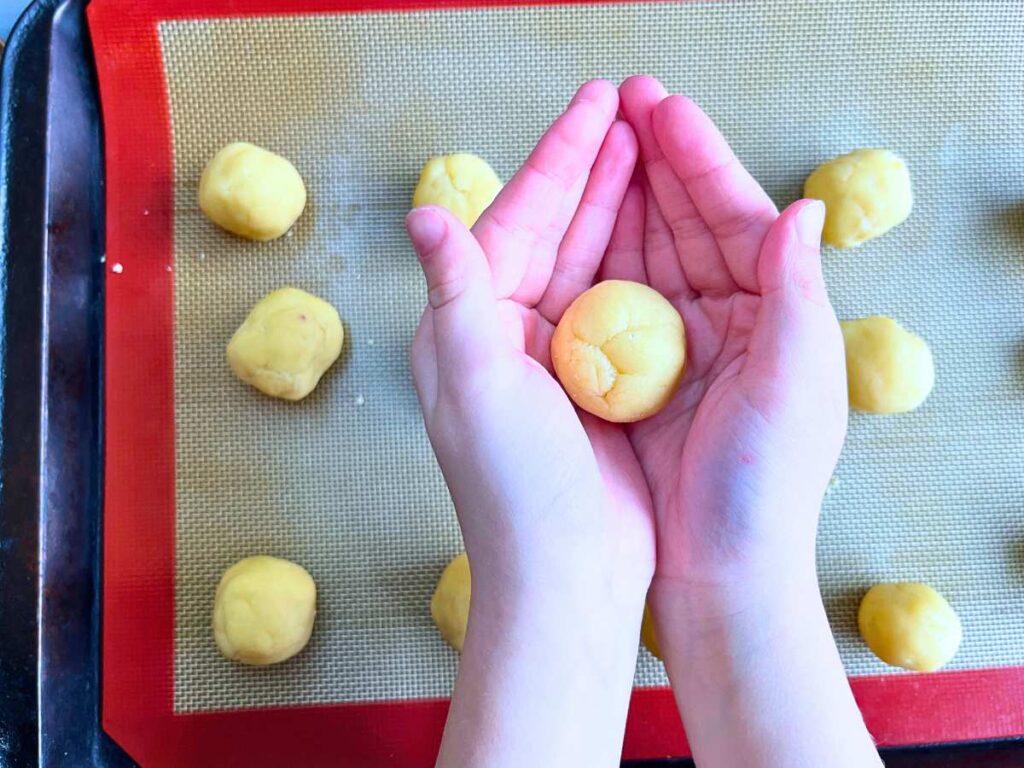 A girl is holding a ball of dough. There is more on a tray.