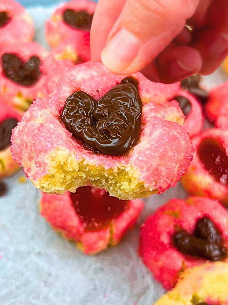 A woman is holding a pink thumbprint cookie with a chocolate heart centre. There is a bite out of it.