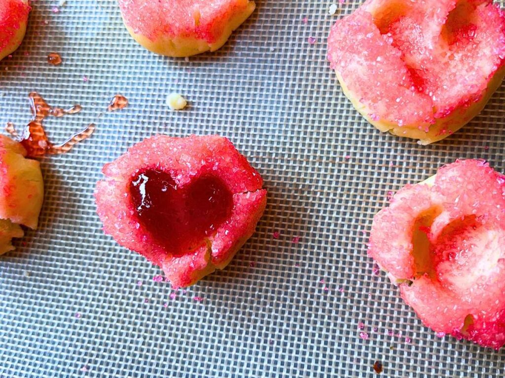 A lined cookie sheet with pink thumbprint cookies with a heart depression in each. Some are filled with jam.