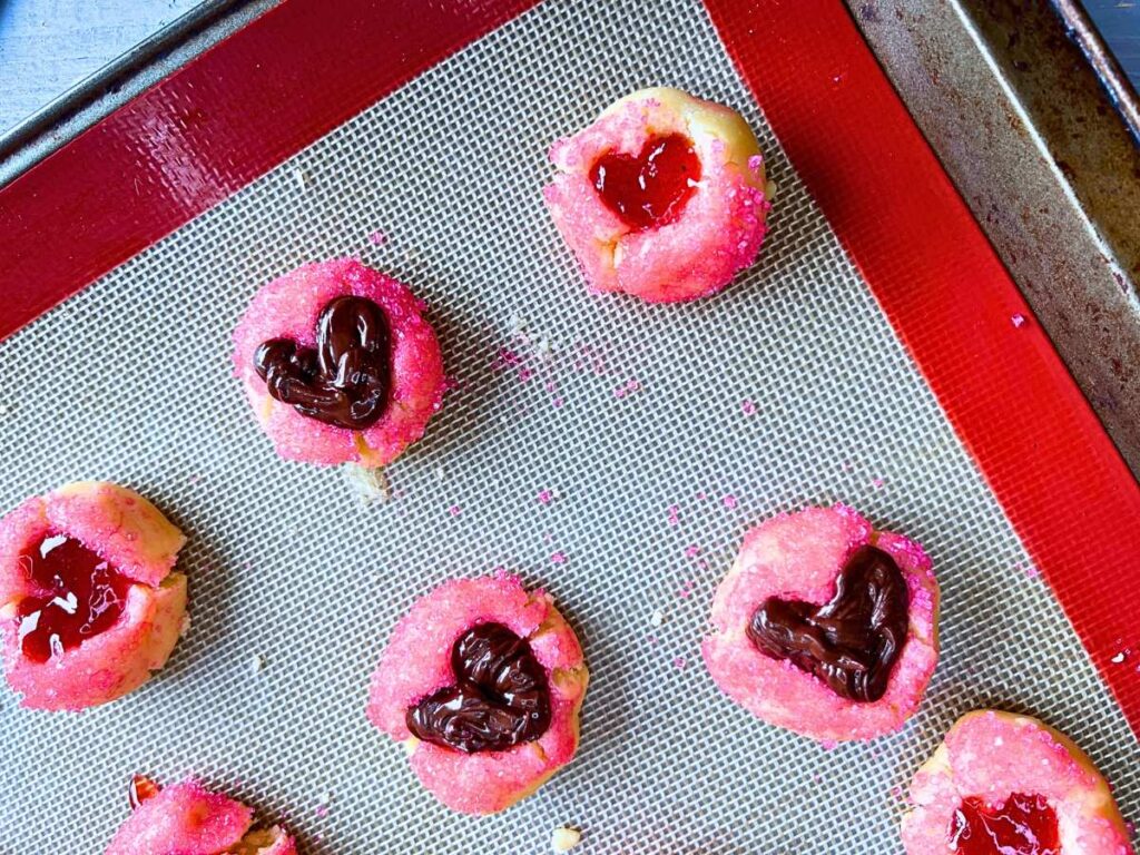 A lined baking sheet with pink thumbprint cookie dough. There are heart depressions in each. Some are filled with jam and some with melted chocolate.