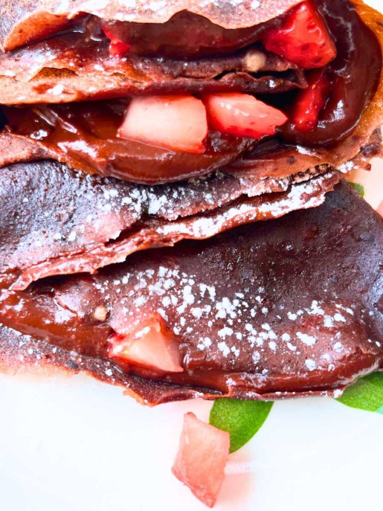 Some folded chocolate crêpes on a plate. They are full of chocolate and diced strawberries.