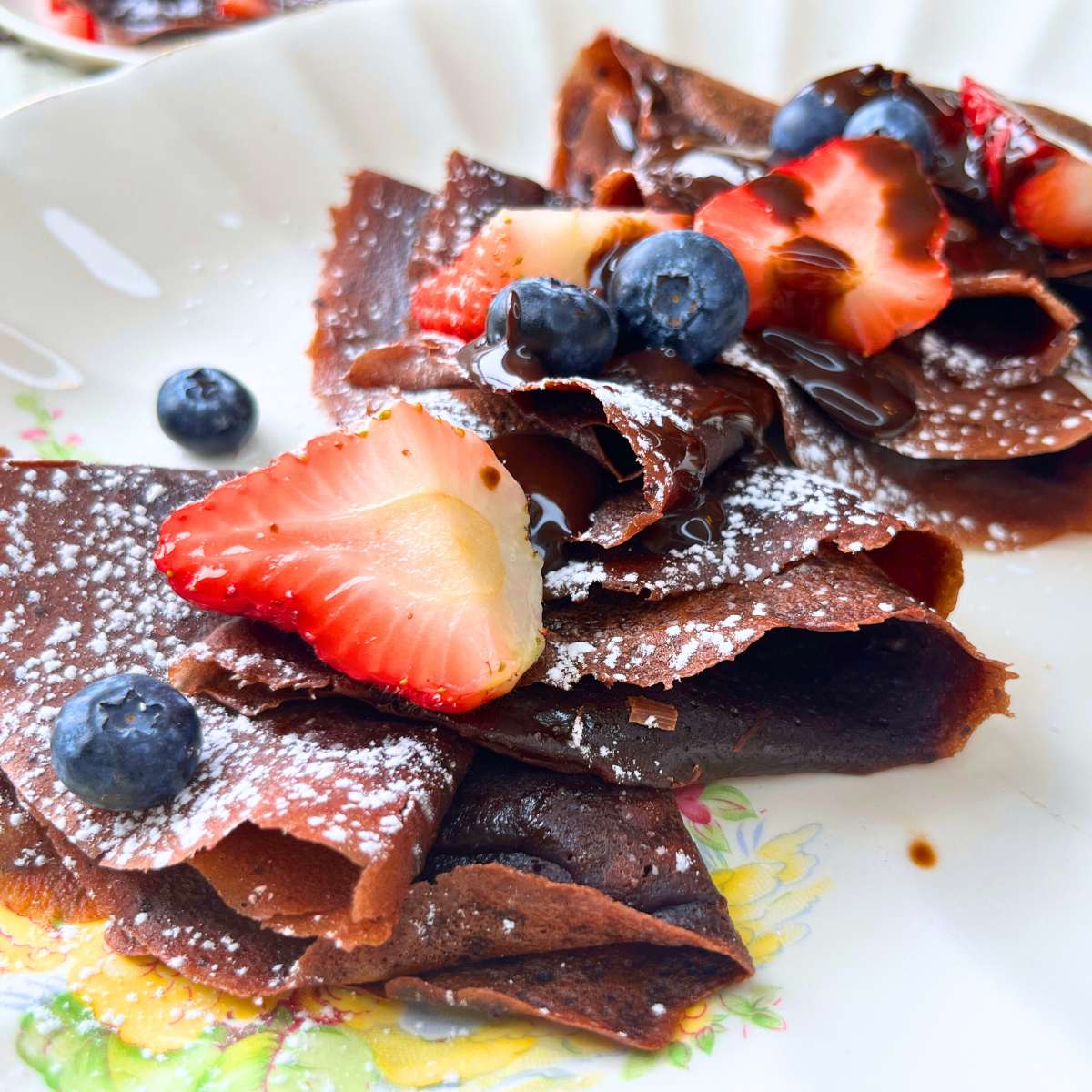 A white plater with folded chocolate crêpes and fresh fruit on top.