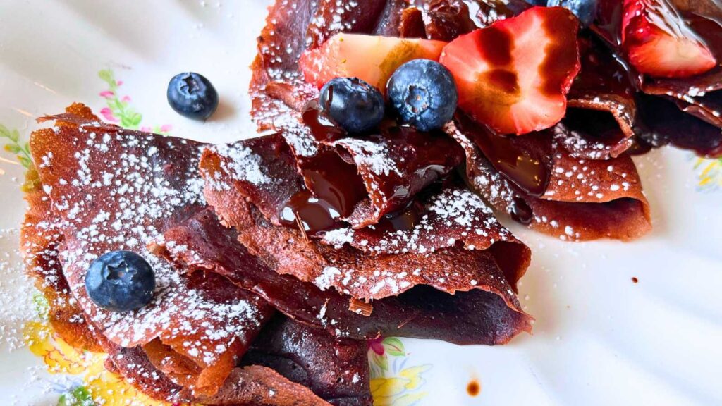 A platter of folded chocolate French crêpes covered in fresh fruit, powdered sugar, and chocolate sauce.