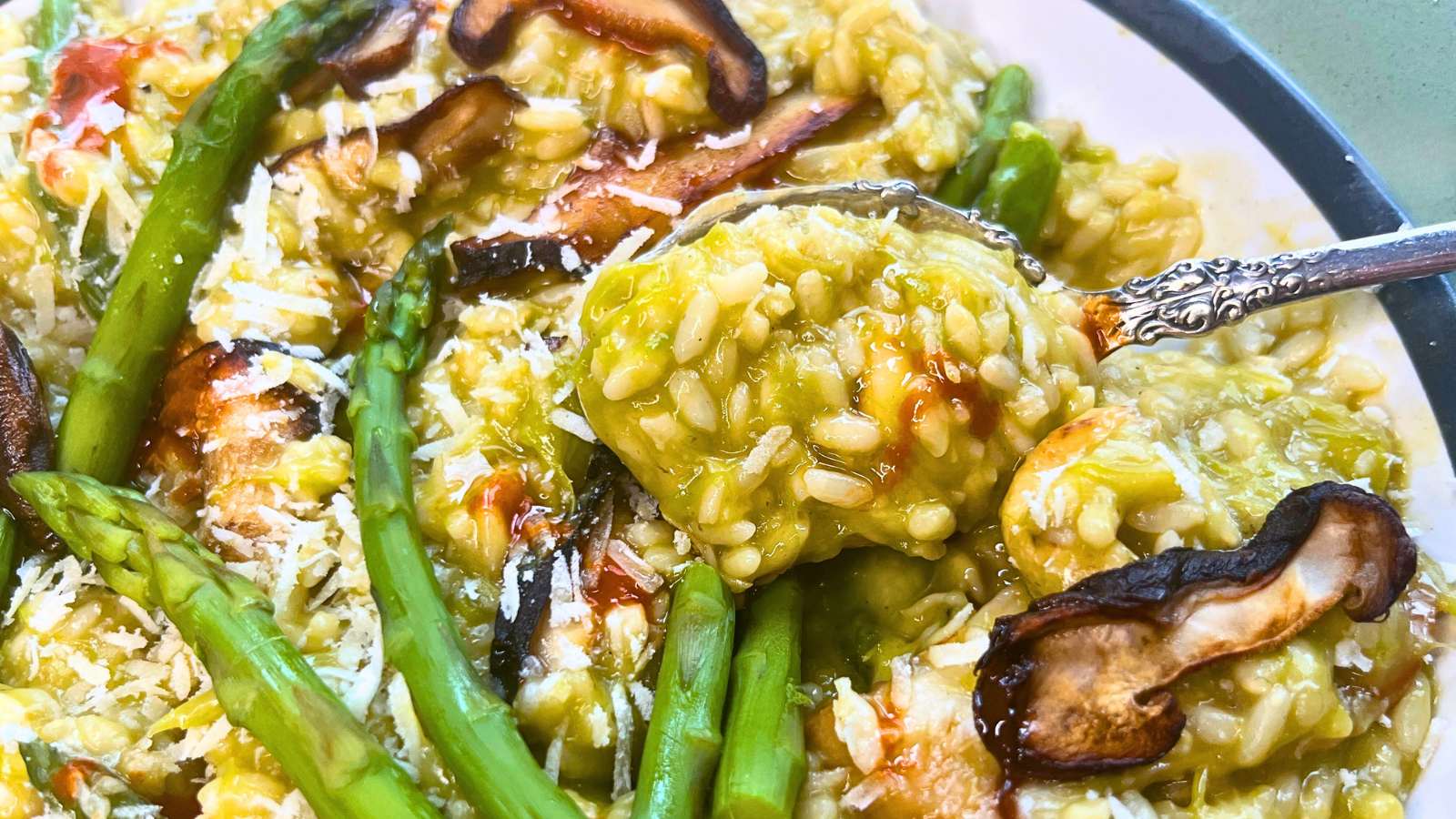 A large white and green bowl filled with risotto. It is covered with cooked asparagus, mushrooms, and cashews. A woman is holding a spoonful of the risotto.