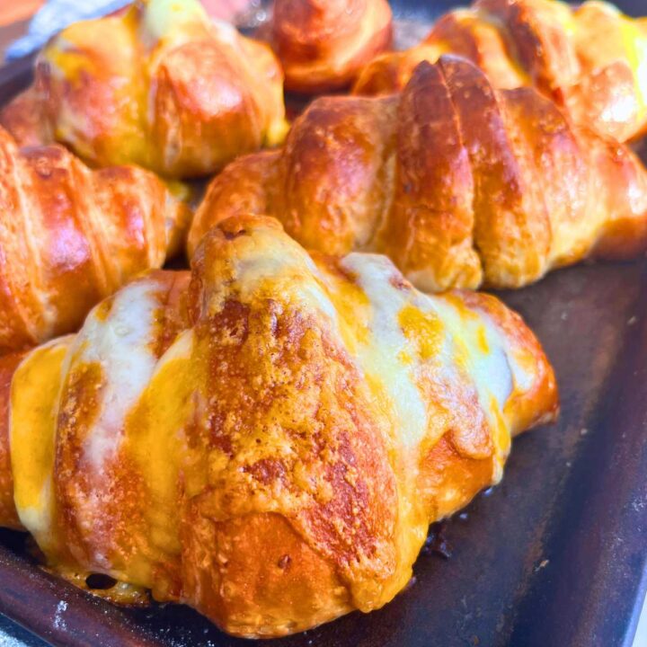 A group of croissants on a baking sheet. Some have melted cheese on top.