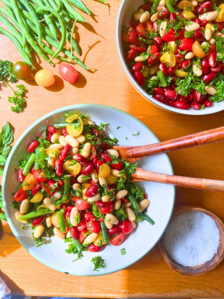 A large white bowl with a bean and tomato salad. There are wooden salad forks inside. There is another bowl of salad behind.