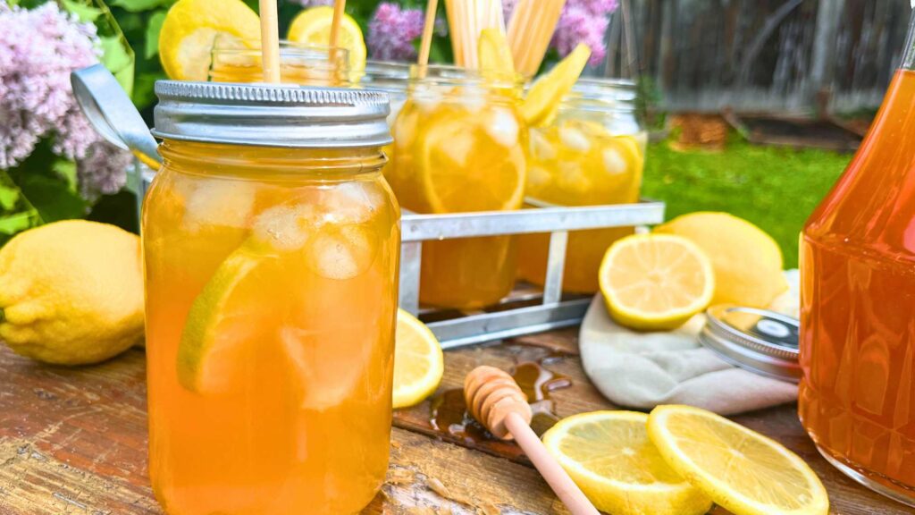 A side view of a glass jar cup with iced tea inside. There is ice and lemon slice in the cup. There are more cups of iced tea in the background.