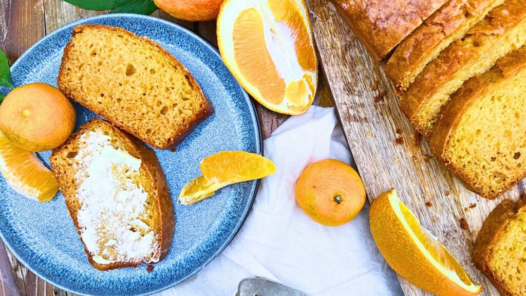 An orange loaf that is partially sliced. Beside is a blue plate with two slices on it. One is buttered.