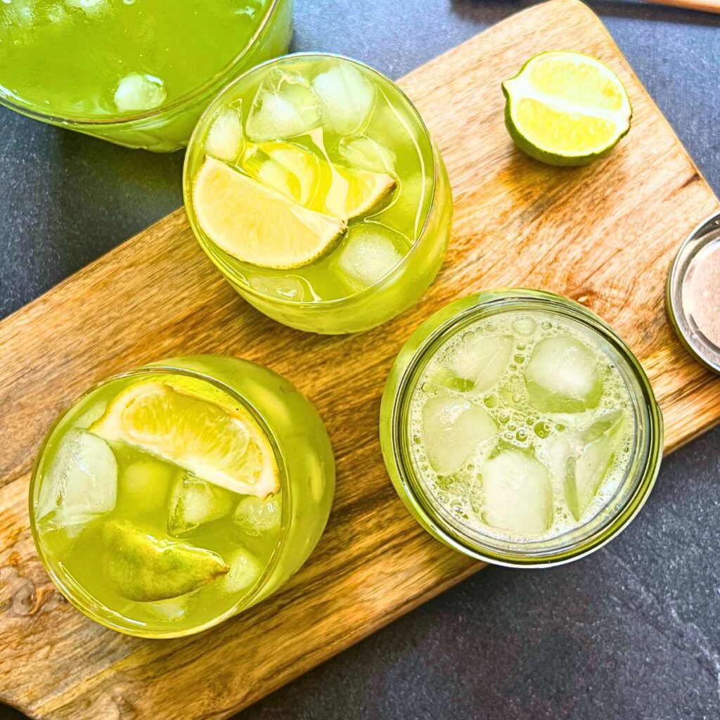 Three glasses full of limeade, lime wedges, and ice. There is a pitcher with more limeade in the background.