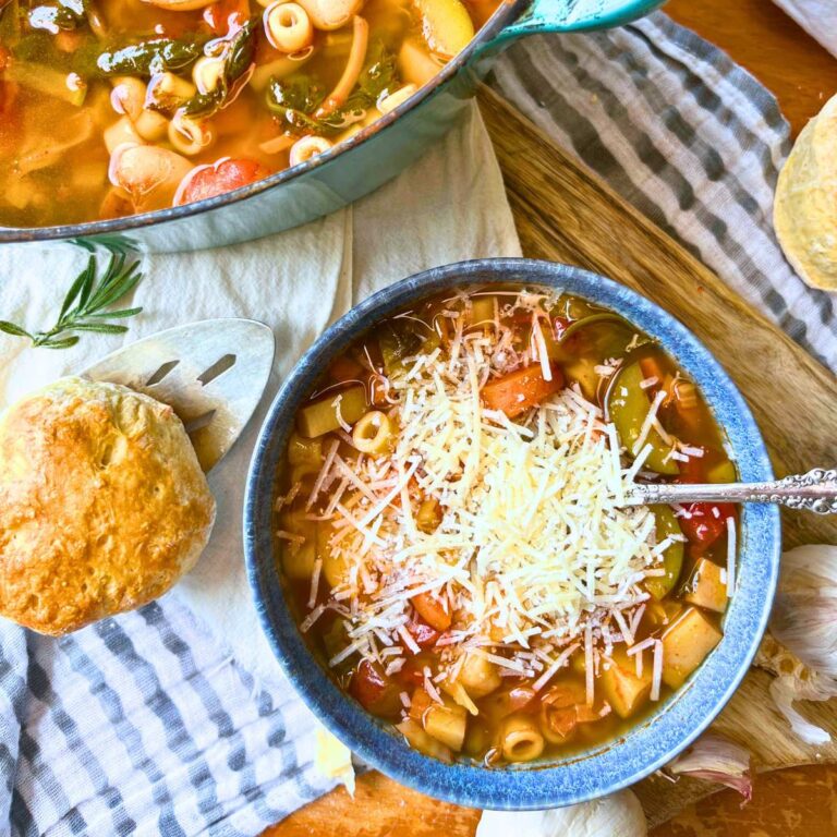 A blue bowl full of minestrone soup with cheese on top. There is more in a pot behind.