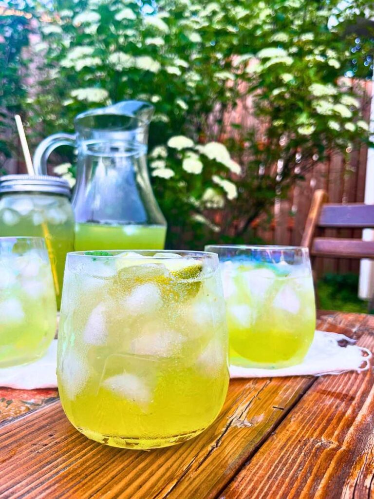 A wooden table with glasses of limeade and ice. There is more in a pitcher in the background.