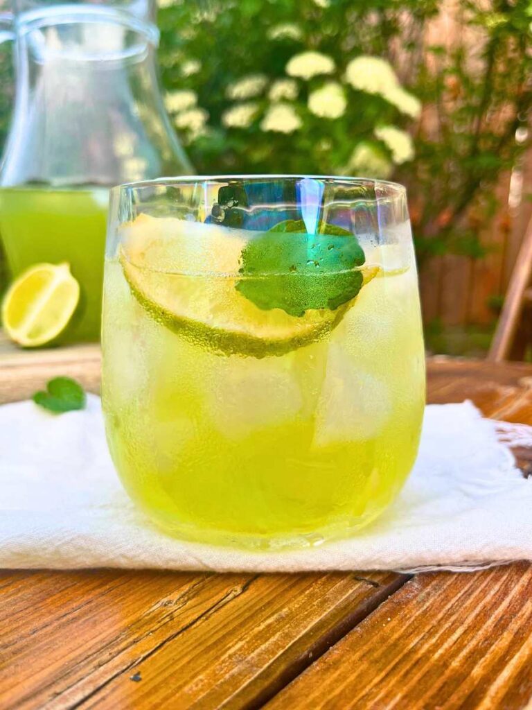A close up look at a glass of limeade with ice. There is a lime wedge and a mint leaf inside. There is more in a pitcher in the background.