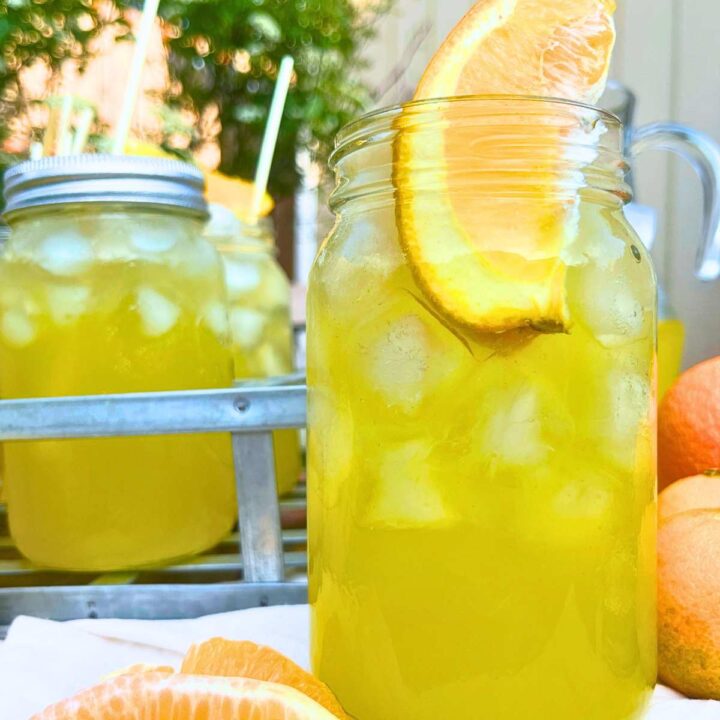 A side view of a glass jar filled with orangeade, ice, and an orange wedge. There is more in the background.