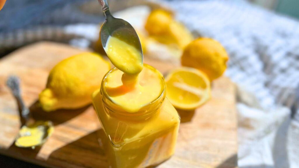 A woman is scooping some lemon curd with a spoon from a glass jar.