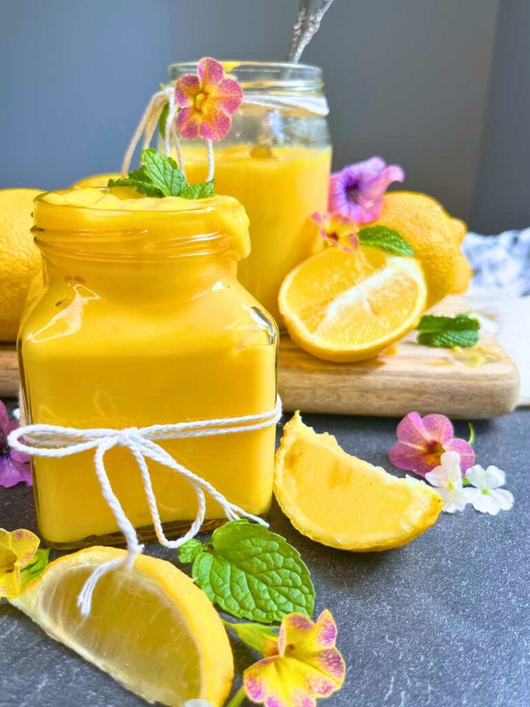 Two jars of eggless lemon curd surrounded by sliced lemons, mint and purples flowers.