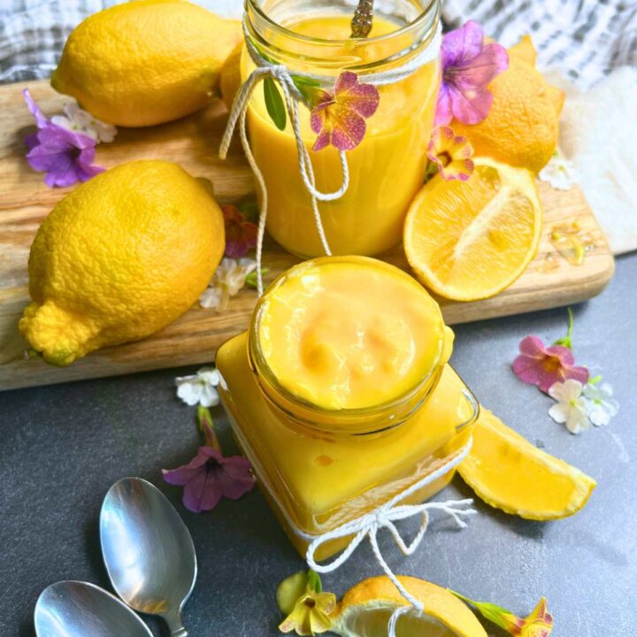 Two jars of homemade lemon curd on a table surrounded by fresh lemons and flowers.