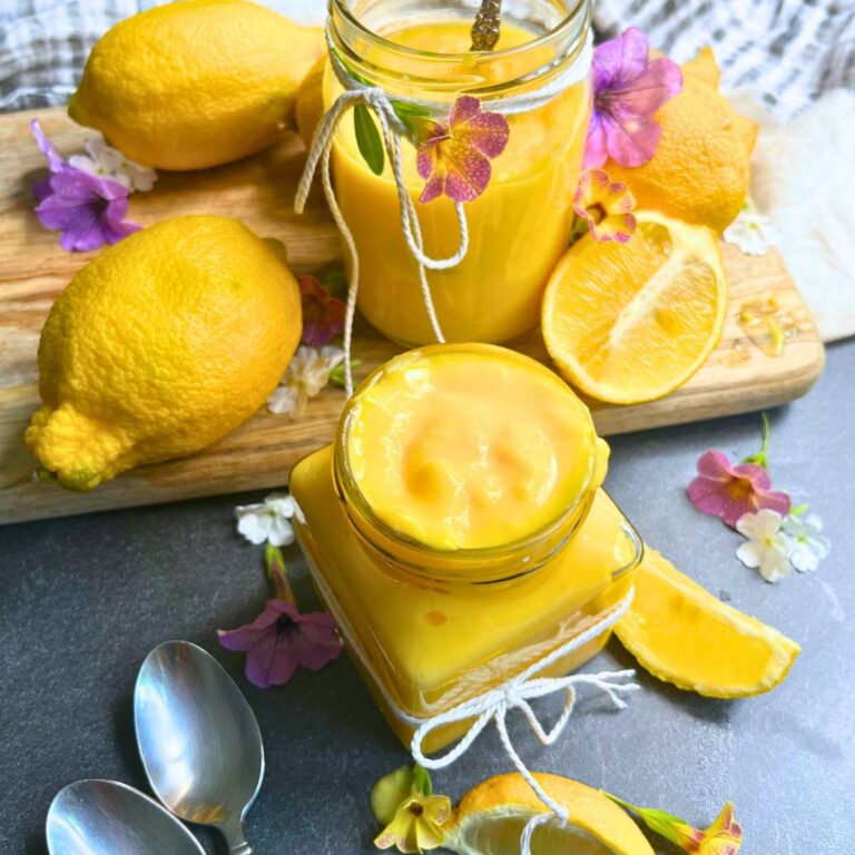 Two jars of homemade lemon curd on a table surrounded by fresh lemons and flowers.
