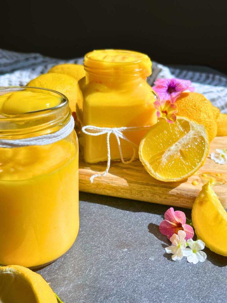 Two glass jars of eggless lemon curd. There are fresh lemons and flowers around the jars.