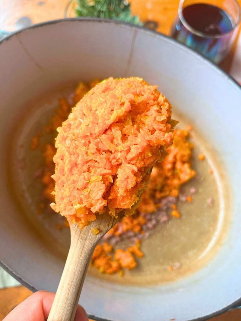 A woman holding up a wooden spoon with a mixture of carrot, celery, and shallot.