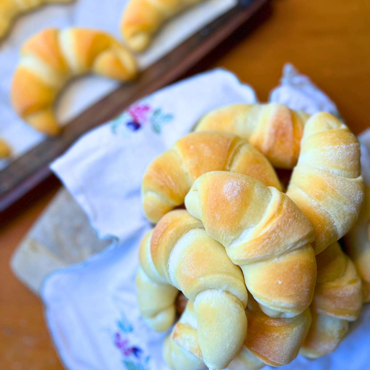 A linen lined basket of crescent rolls. There are more in the background on a baking sheet.