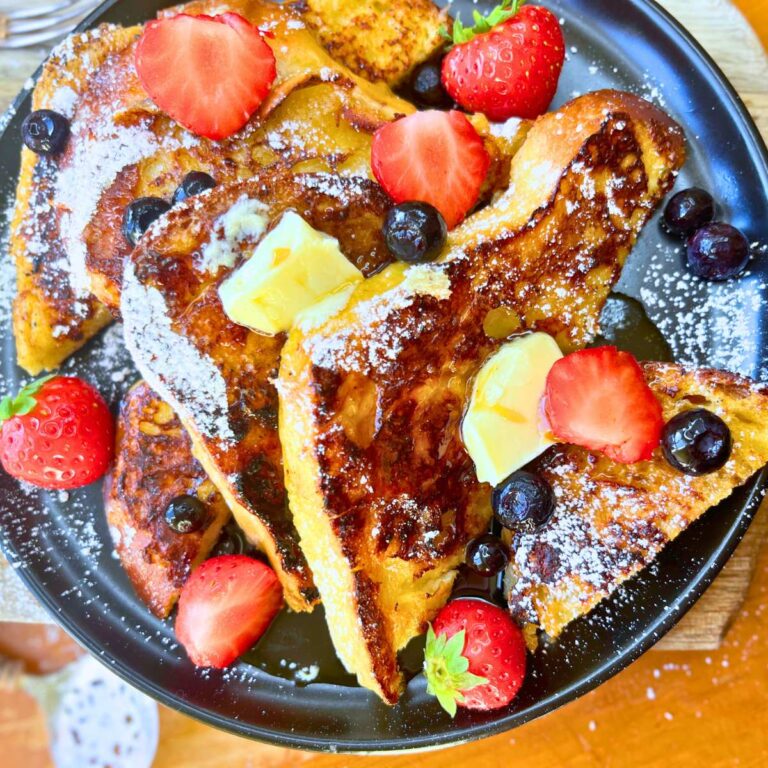 A black plate with sliced French toast covered in sliced strawberries, blueberries, powdered sugar, and syrup.