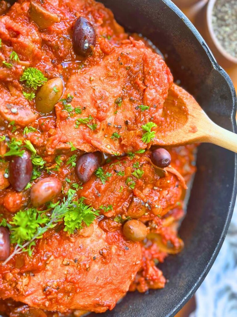 A close up look at a skillet of chicken cacciatore. There is a wooden spoon under one piece of chicken.