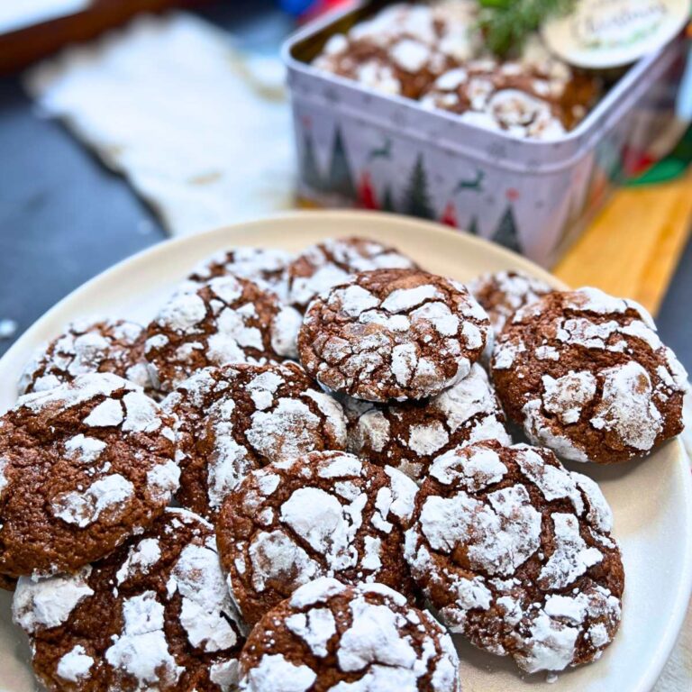 A white plate full of chocolate cookies with a crackled white icing sugar design. There is more in the background in a Christmas tin.