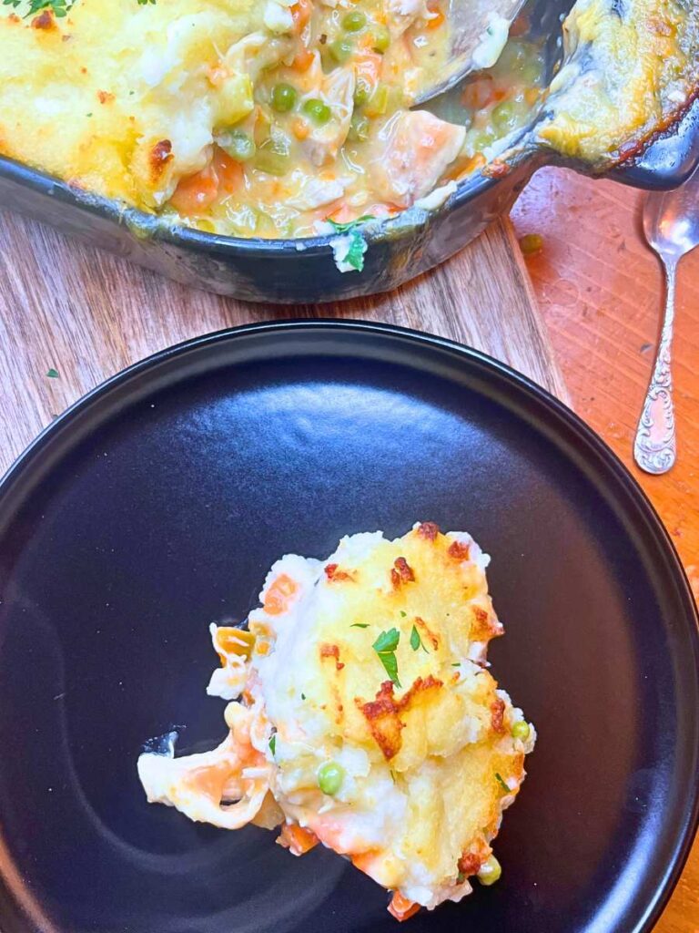 An overhead view of a black casserole dish with a turkey shepherd's pie inside. There is a serving in front on a black plate.