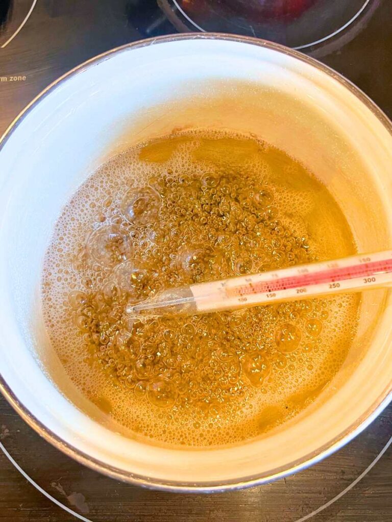 A white pot with cooking sugar inside. There is a candy thermometer in the sugar liquid.