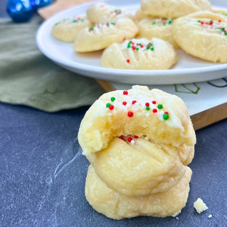 A stack of three whipped shortbread cookies. The one on top has a bite out of it. There are more on a white plate in the background.
