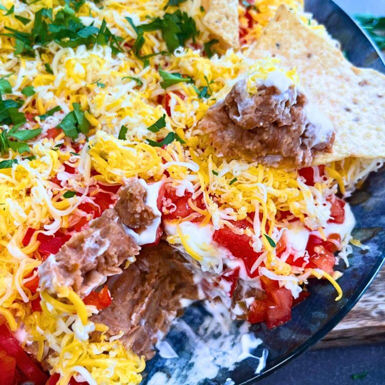 A layered bean dip with a scoop removed. There is a tortilla chip on top.