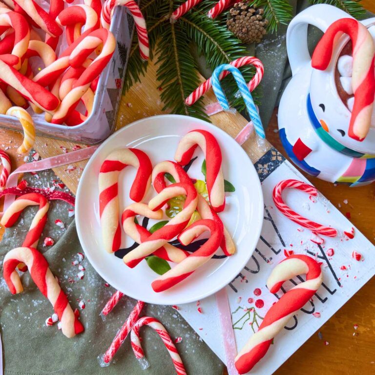 Candy cane cookies on a white plate. There are more to the side in a Christmas tin. There is a mug off to the side.
