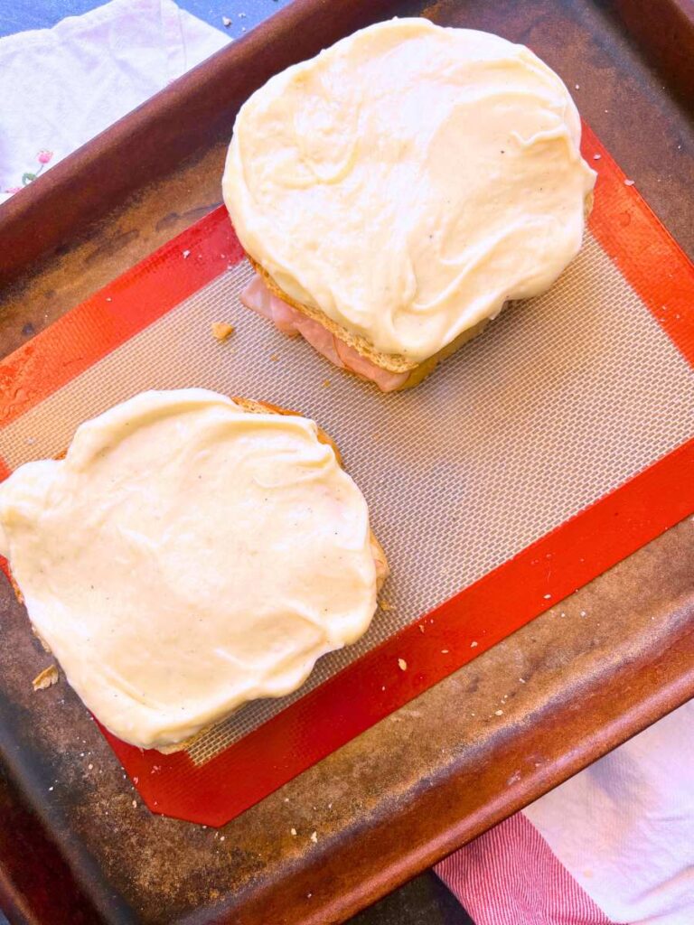 A baking sheet with two ham and cheese sandwiches with mornay sauce on top.