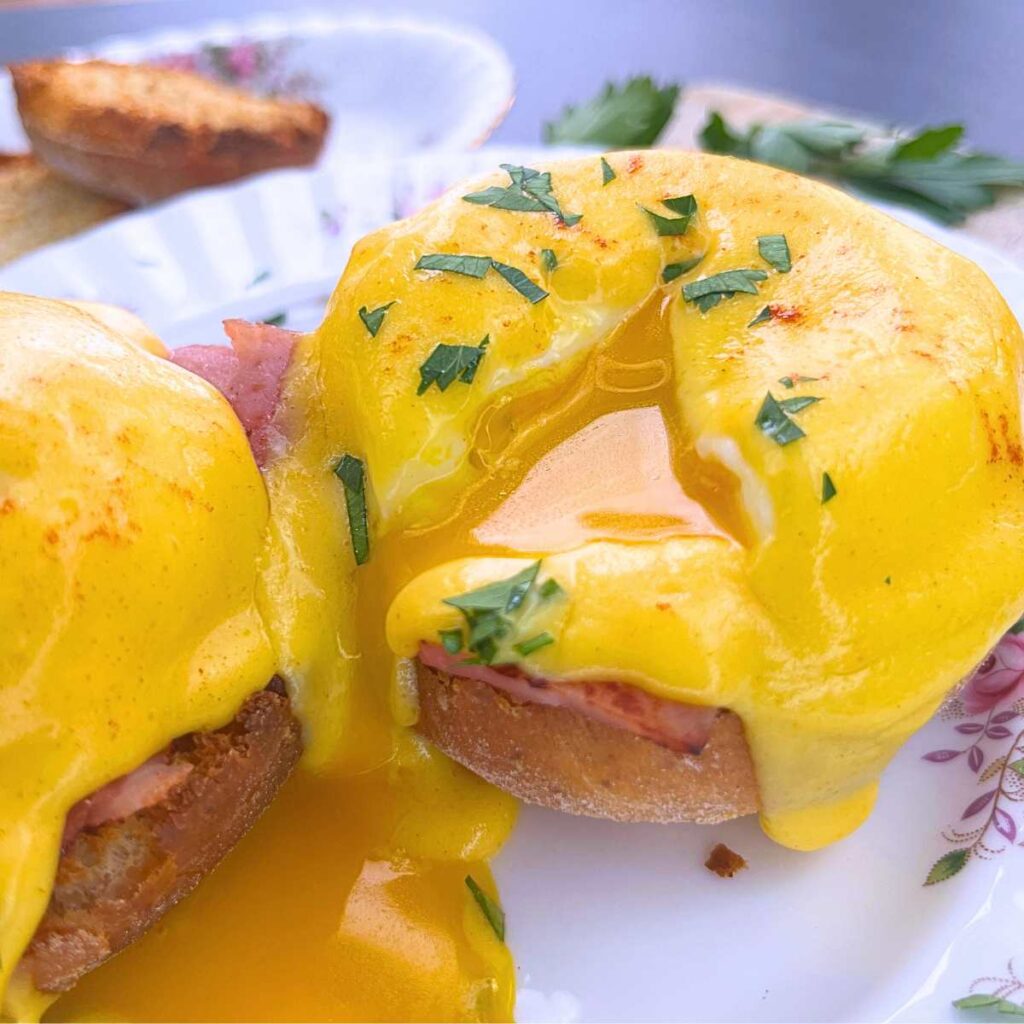 Two eggs Benedict on a pink floral plate. Once has a slice in the poached egg to show the runny yolk inside.